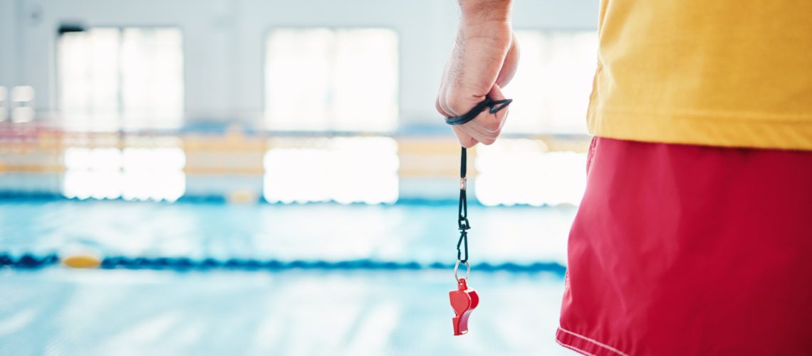 Hands, lifeguard and whistle by swimming pool for water safety, security or ready for rescue indoors. Hand of expert swimmer holding signal tool for warning, safe swim or responsibility for awareness
